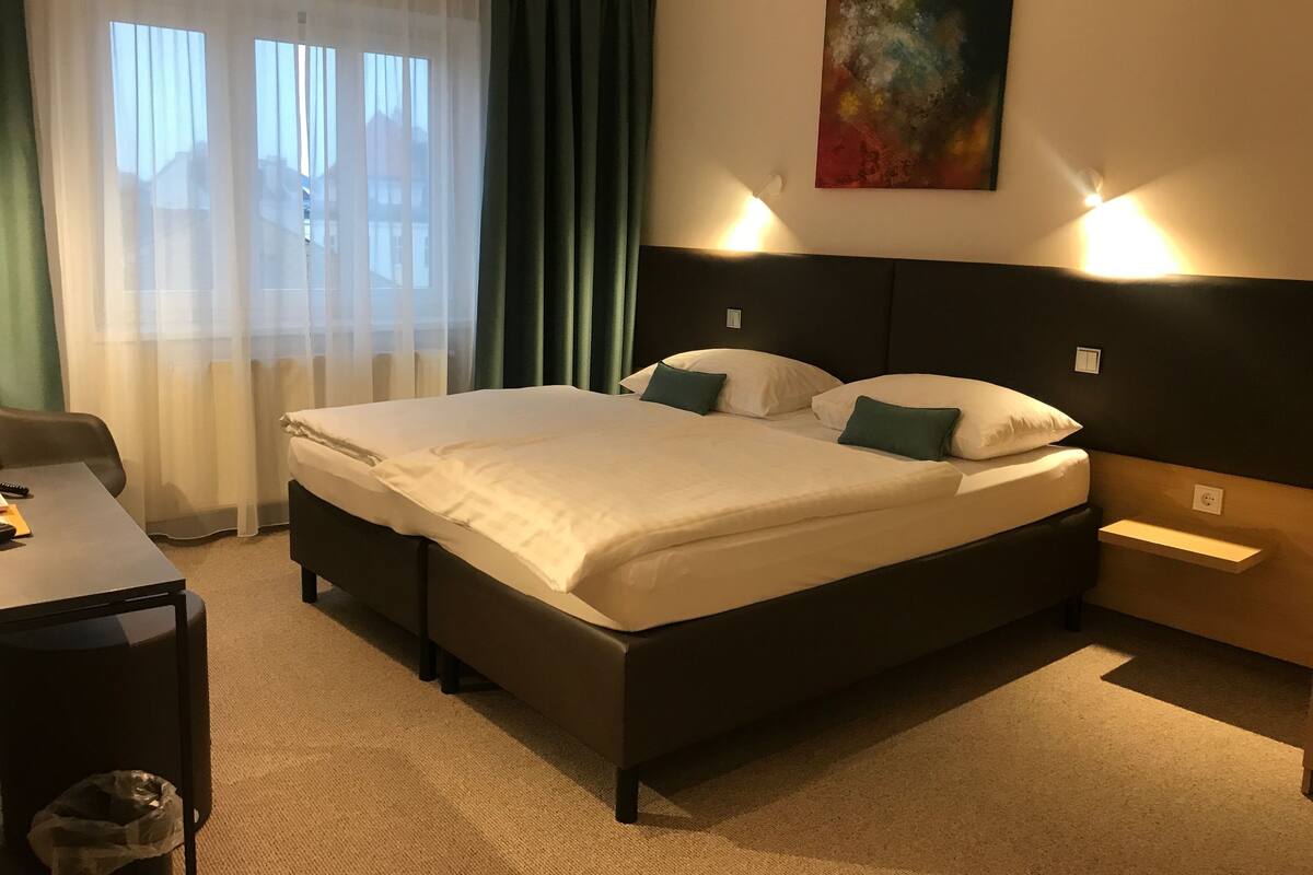 A hotel room with a large bed, a desk, a chair, a trash bin, and a painting on the wall.