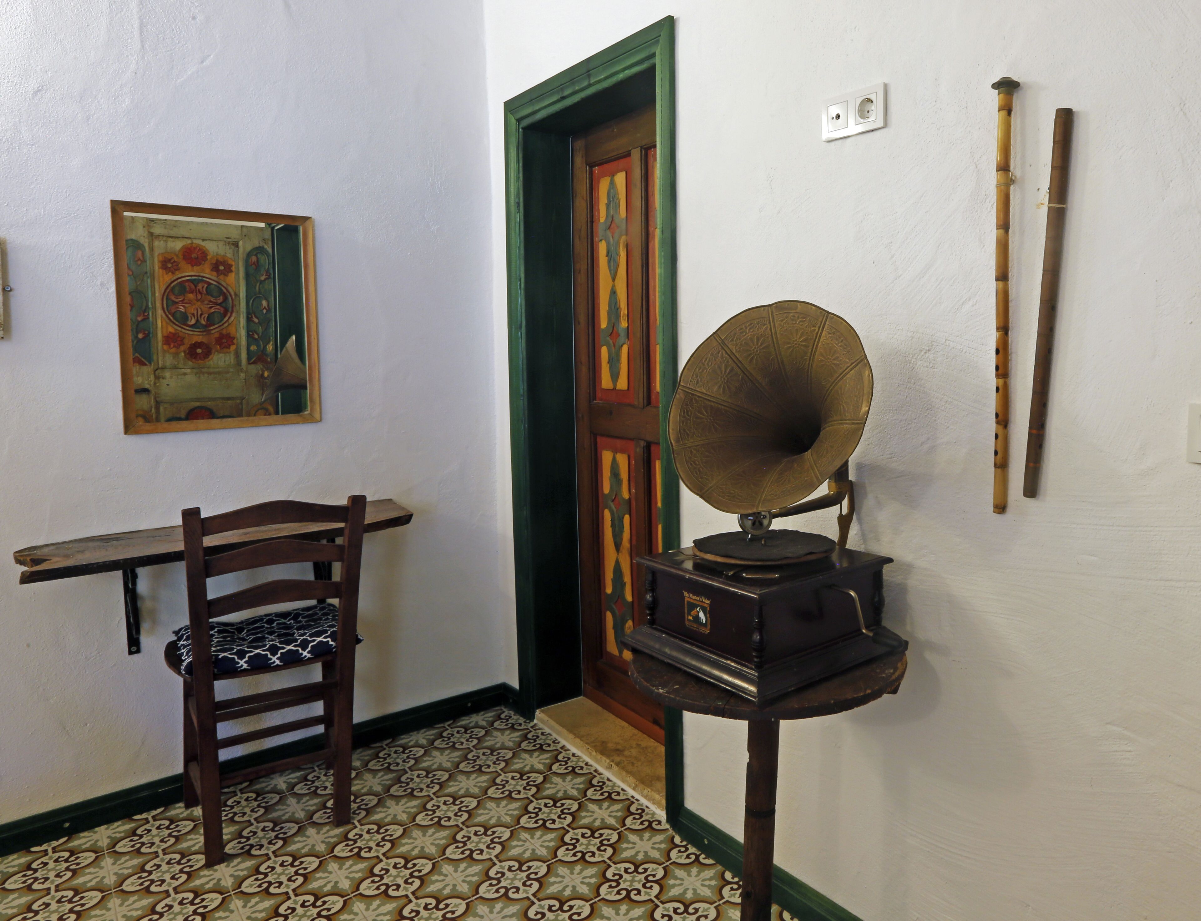 A room with a wooden chair, a gramophone, a patterned floor, a door, and a wall-mounted picture.