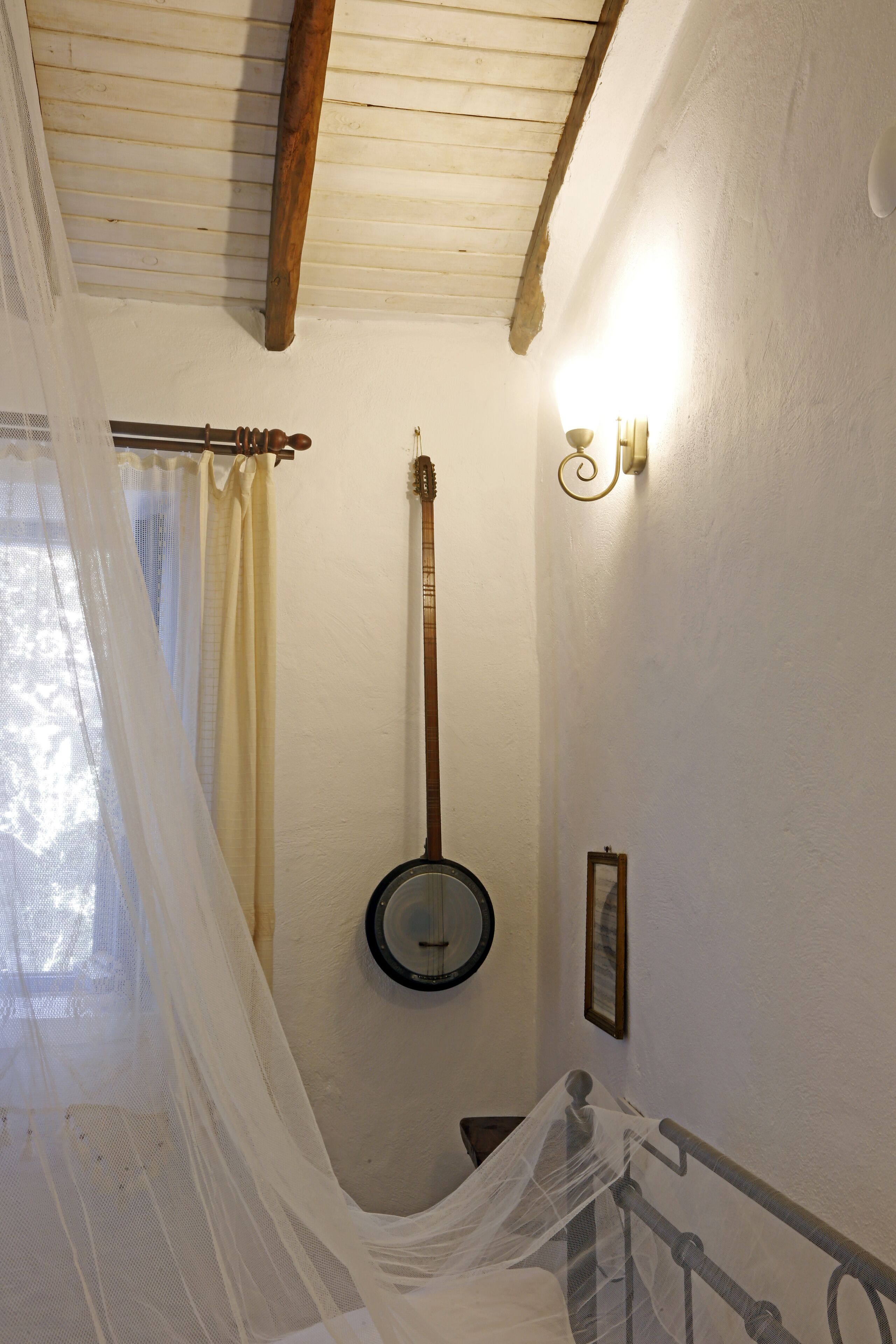 A room with a wooden ceiling, a banjo hanging on the wall, a bed with a white bedspread, and a window with a sheer curtain.
