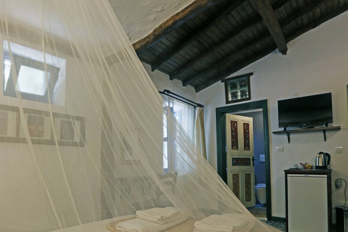 A bedroom with a bed covered by a mosquito net, a small table with folded towels, a television mounted on the wall, and a door leading to another room.