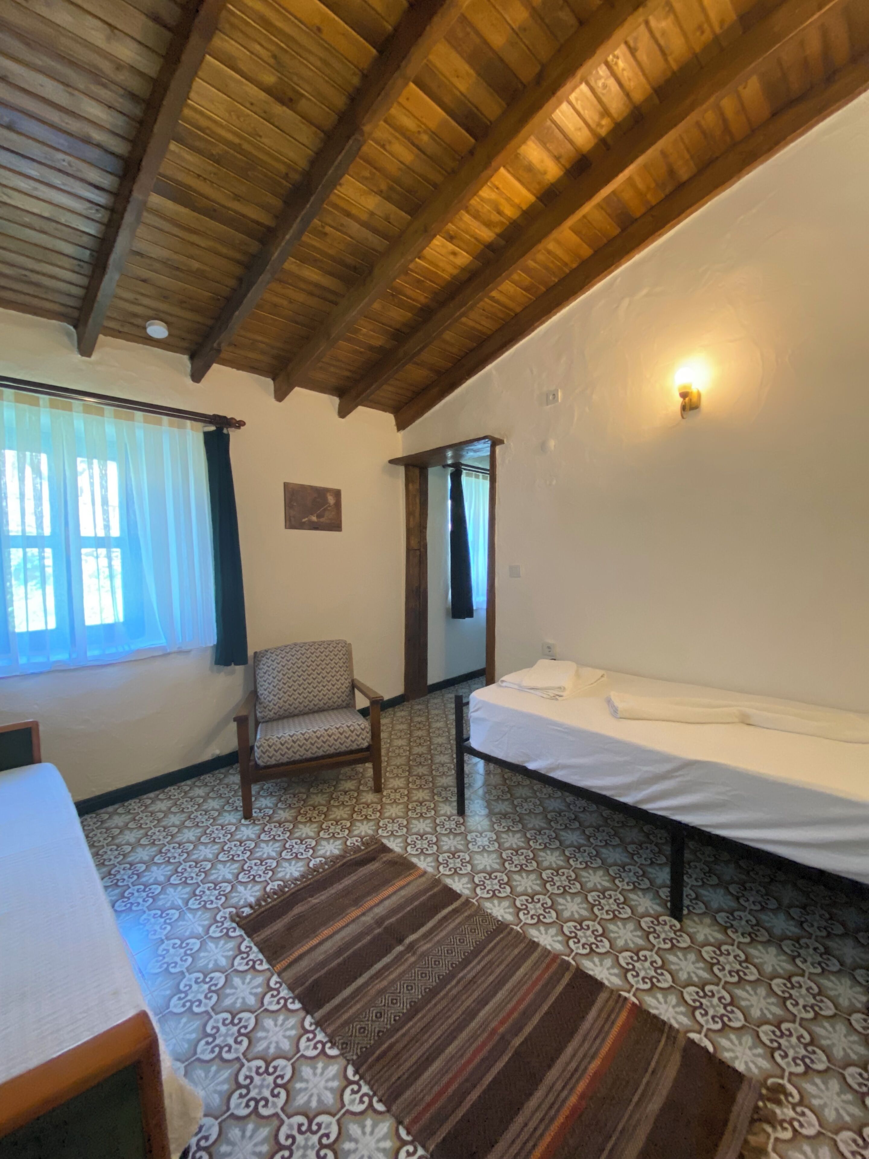 A room with a bed, a chair, a window with curtains, a patterned floor, and a wooden ceiling.