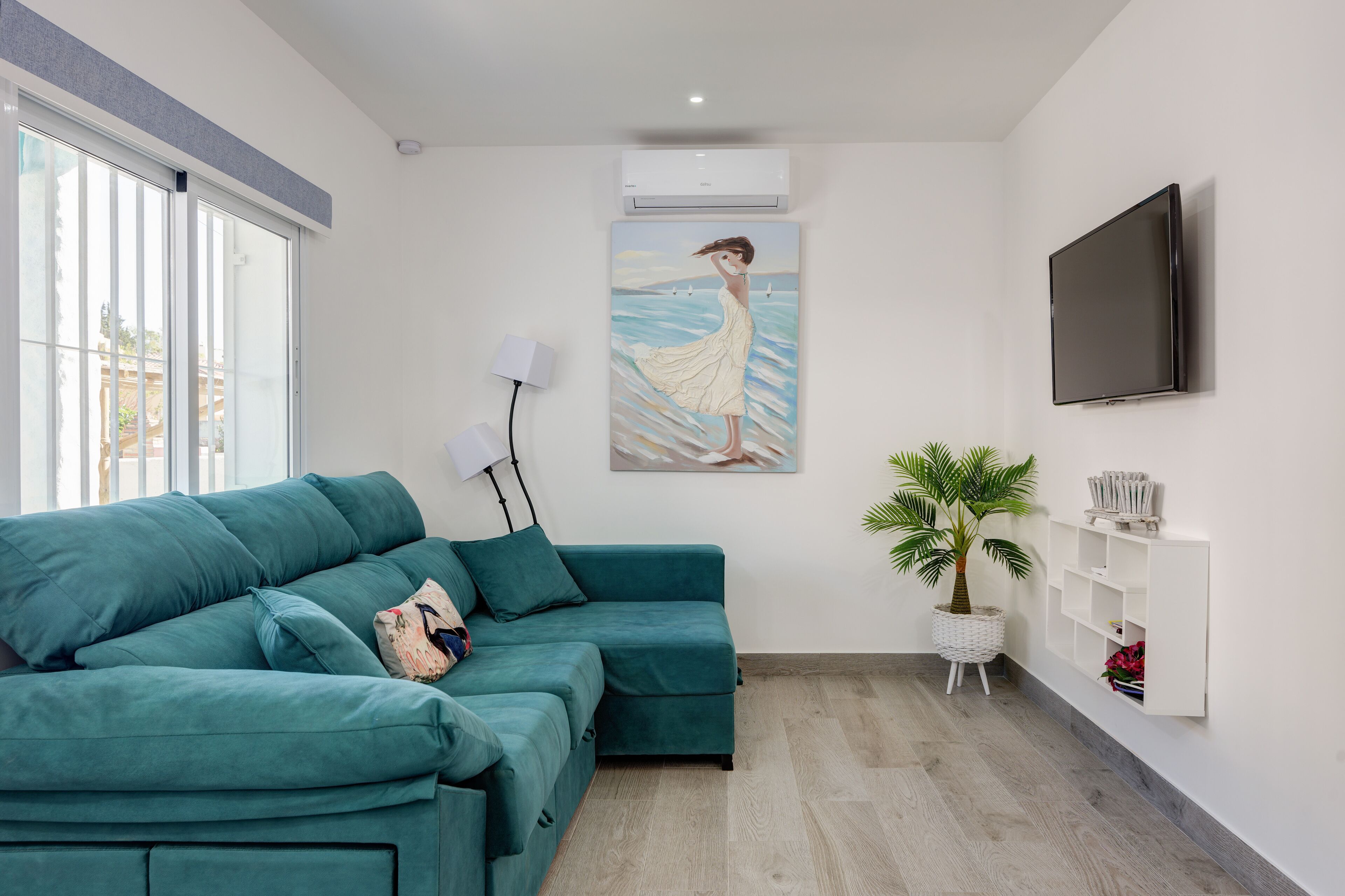 A teal-colored sofa in a room with a large window, a wall-mounted TV, and a painting of a woman by the sea.