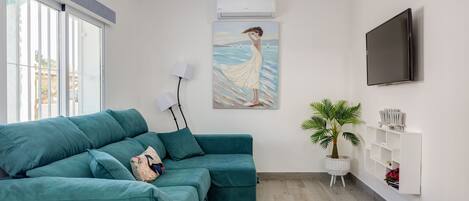 A teal-colored sofa in a room with a large window, a wall-mounted TV, and a painting of a woman by the sea.
