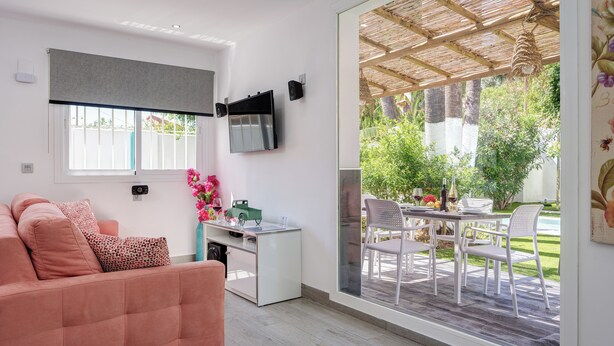 Ein modernes Wohnzimmer mit einer Couch, einem Schreibtisch und einem Fernseher. Ein großes Fenster bietet Blick auf einen Garten mit Gartenmöbeln.