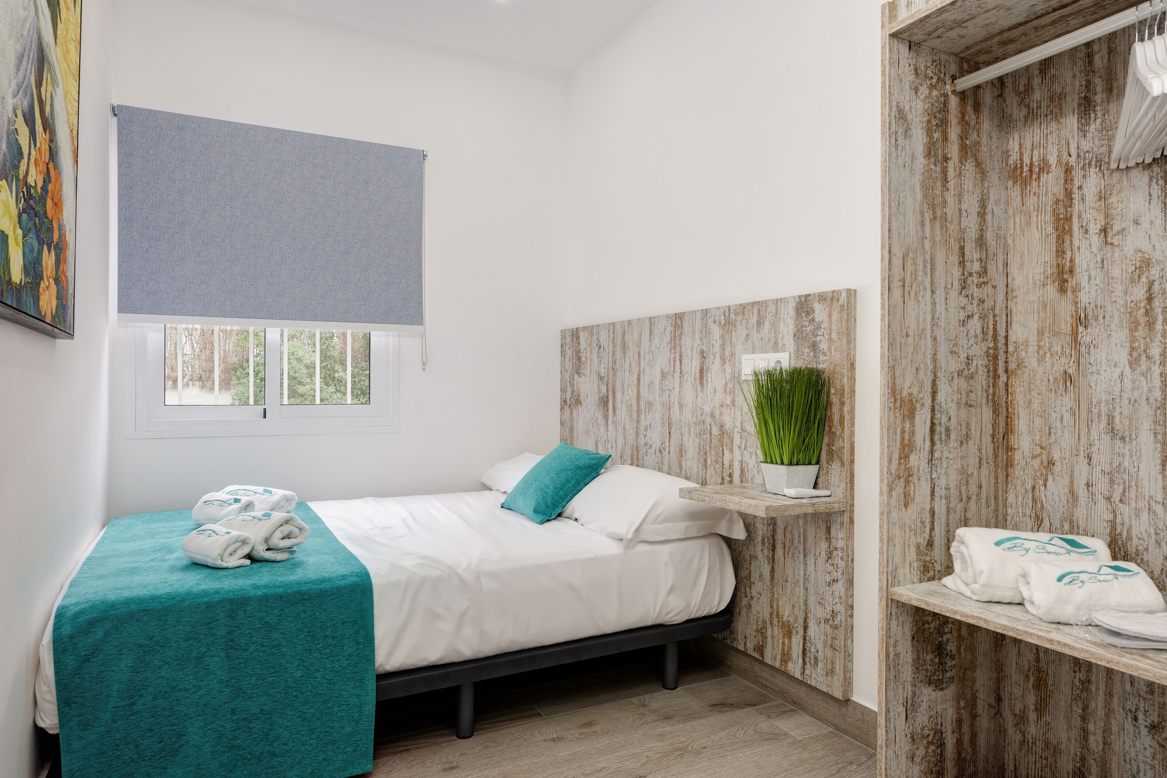 A single bed with a teal bedspread, white linens, and a teal pillow. There is a window with a gray blind, a wooden wall with a distressed finish, and a small shelf with towels and a potted plant.
