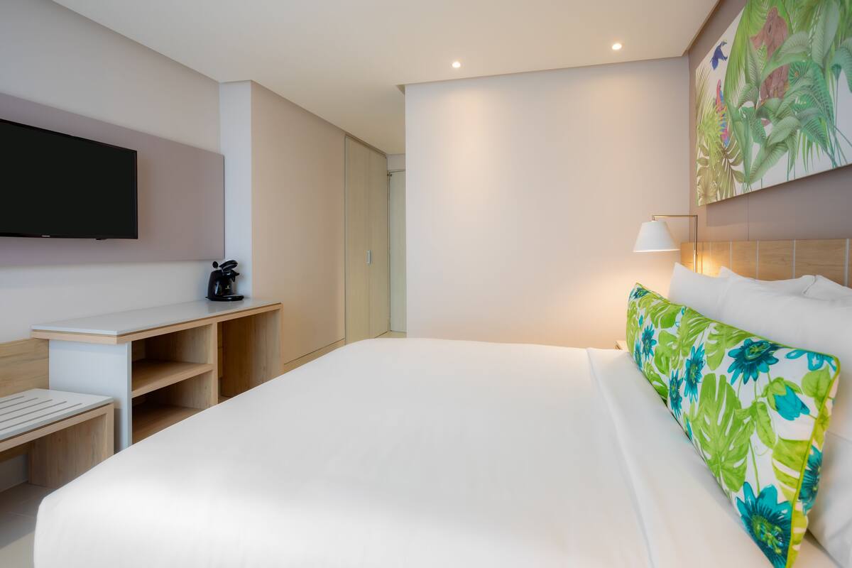 A hotel room with a bed, a desk, a flat-screen TV, a coffee maker, and a wall-mounted lamp.