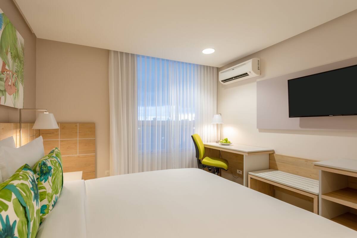 A hotel room with a bed, a desk, a chair, a TV, and an air conditioning unit.
