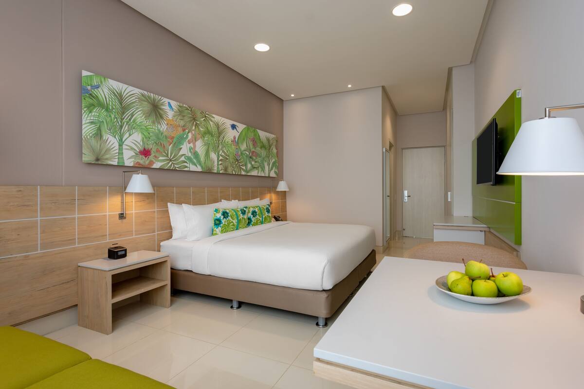 A modern hotel room with a bed, bedside table, lamp, and a fruit bowl on the nightstand.