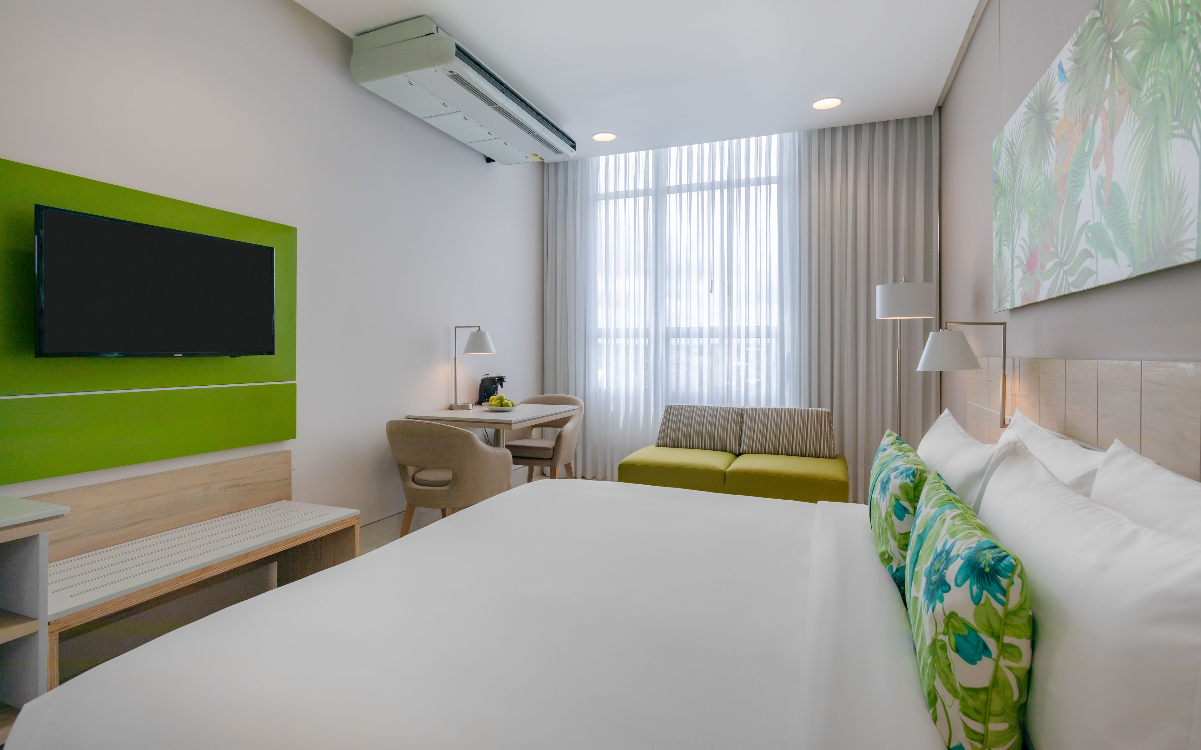 Habitación de hotel con una cama grande, un escritorio, una silla, una mesita, un sofá y un televisor montado en la pared.