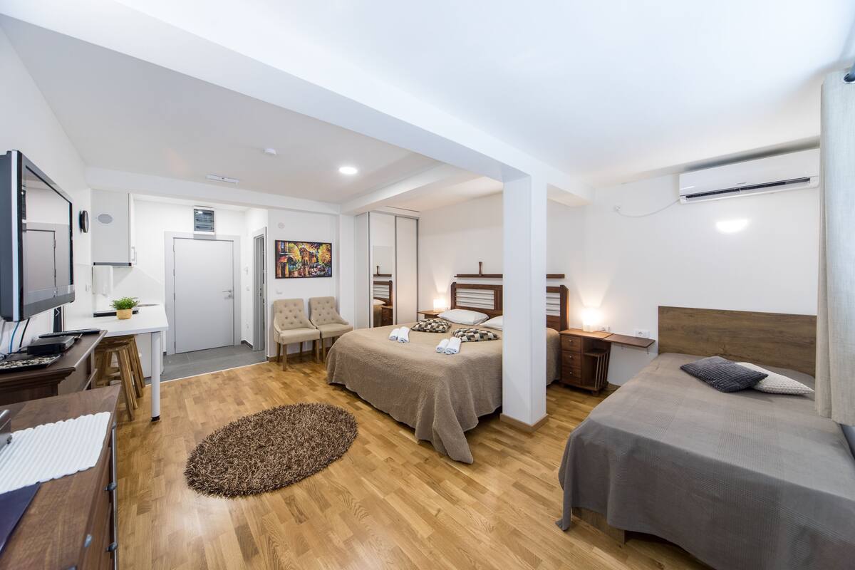 Quarto de hotel moderno com piso de madeira, cama com cabeceira, escrivaninha com computador, cadeira e um quadro na parede.