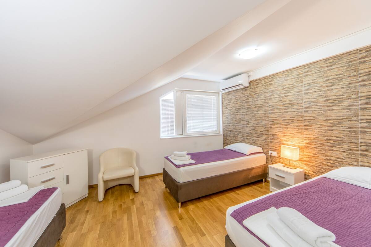 Quarto moderno com duas camas, um guarda-roupa branco, uma cadeira e uma janela com persianas.