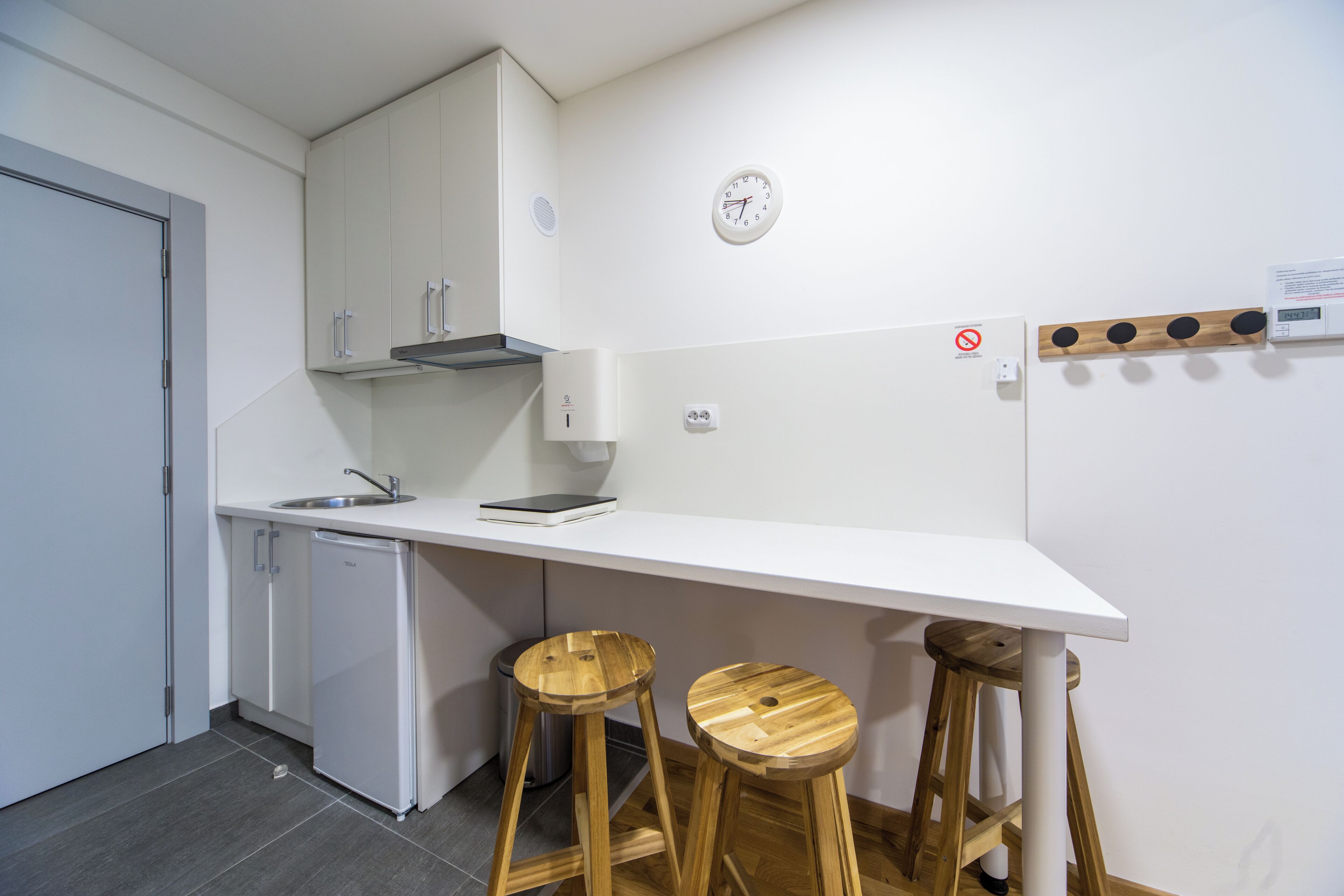 A modern kitchen with white cabinets, a built-in dishwasher, a wooden bar stool, and a wall-mounted clock.