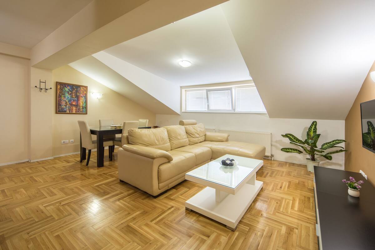 Uma sala de estar moderna com piso em padrão herringbone de madeira, um sofá seccional bege, uma mesa de centro de vidro e uma área de jantar com mesa e cadeiras.