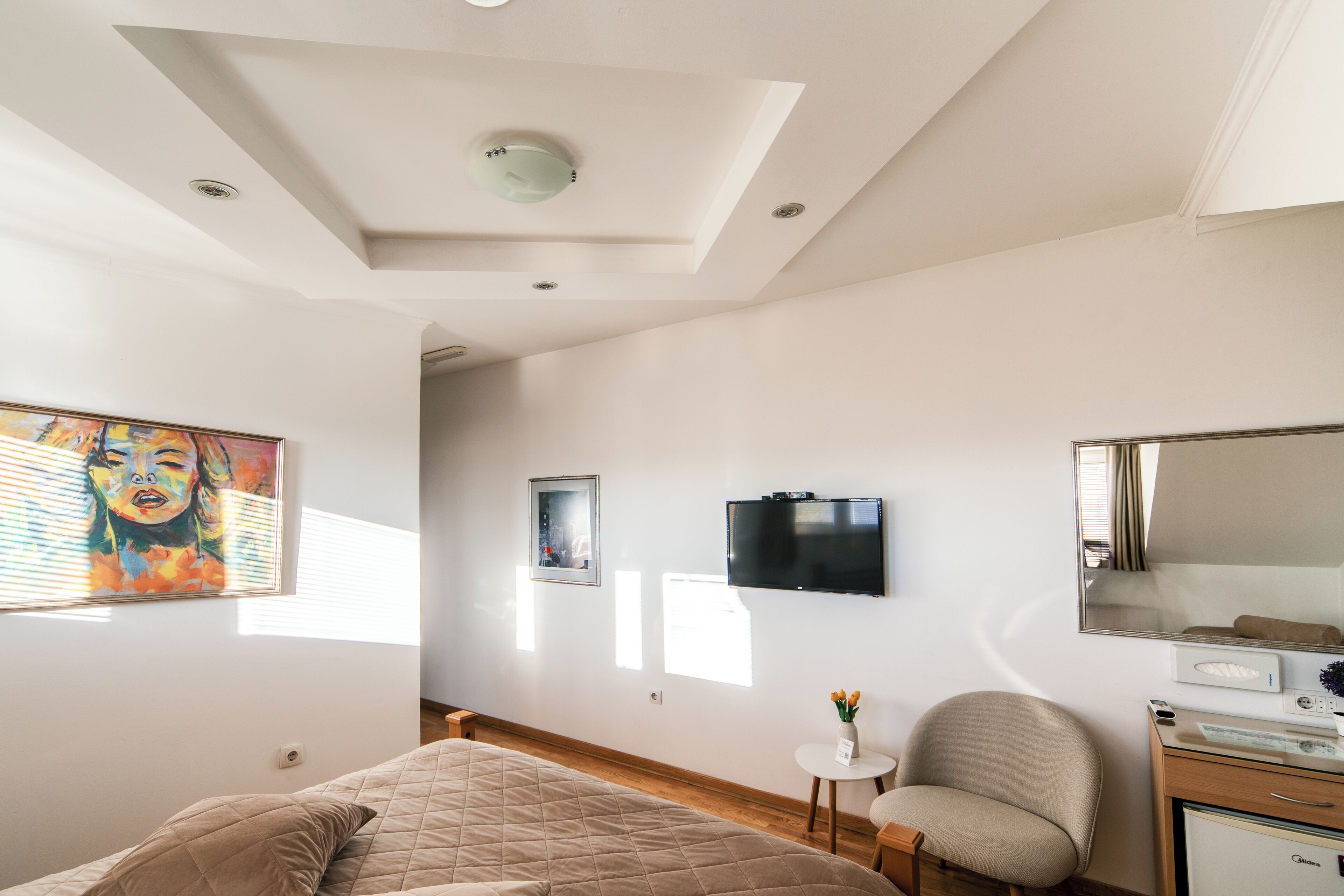 A bedroom with a bed, a chair, a small table with a vase, a TV, and a mirror.