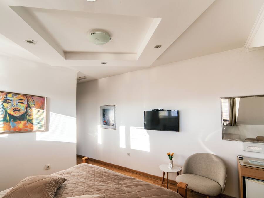 A bedroom with a bed, a chair, a small table with a vase, a TV, and a mirror.
