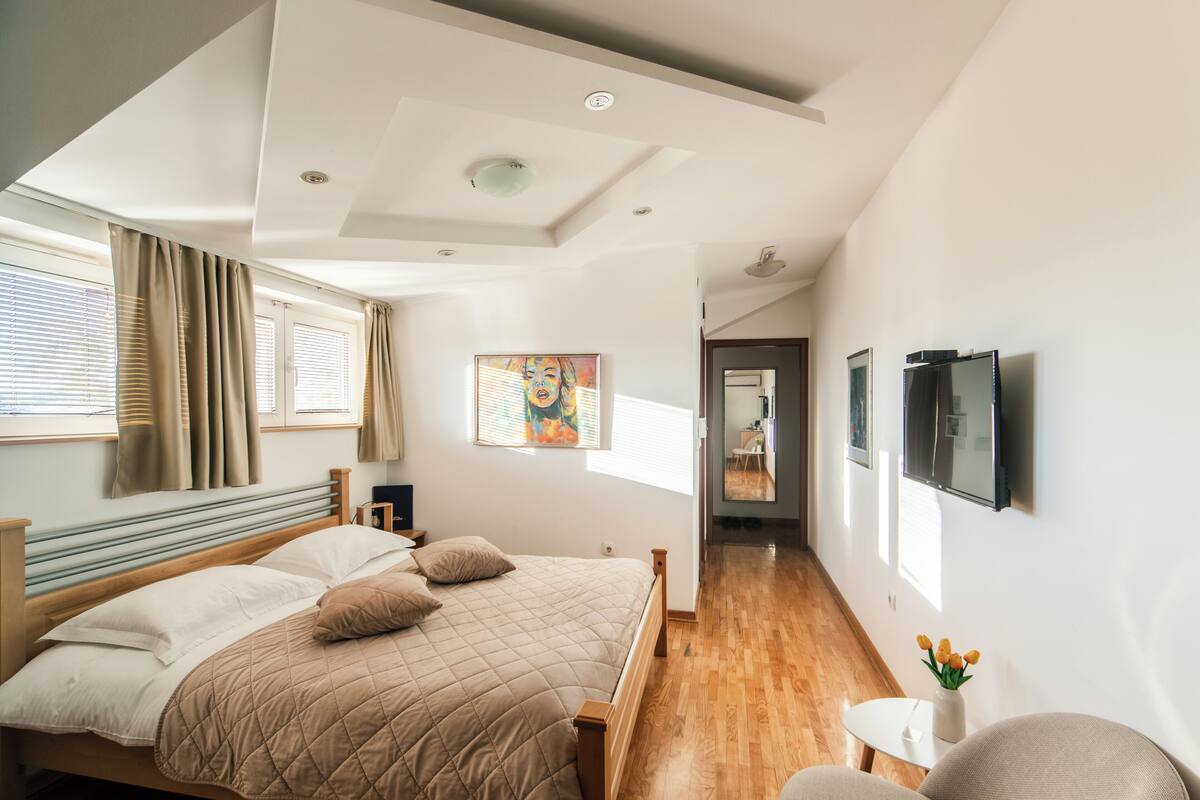 Quarto moderno com uma cama grande, cabeceira de madeira e uma pequena mesa de cabeceira branca com um vaso de flores.