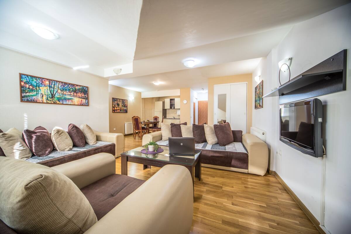 Uma sala de estar moderna com sofás, uma mesa de centro e uma televisão. É visível uma área de jantar ao fundo.