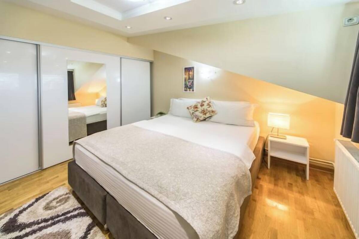 Quarto com cama, mesa de cabeceira, luminária e vista para outro quarto com cama.