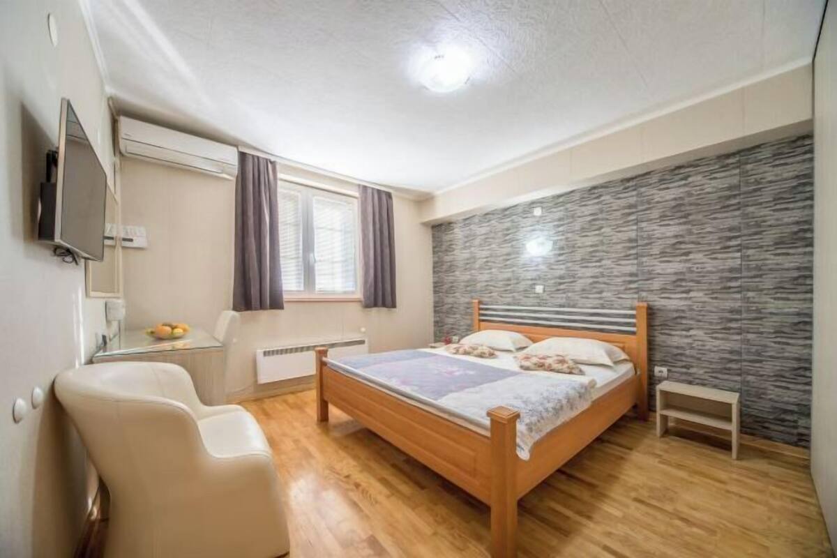 Quarto com cama de madeira, mesa de cabeceira pequena, uma cadeira, televisão e janela com cortinas.