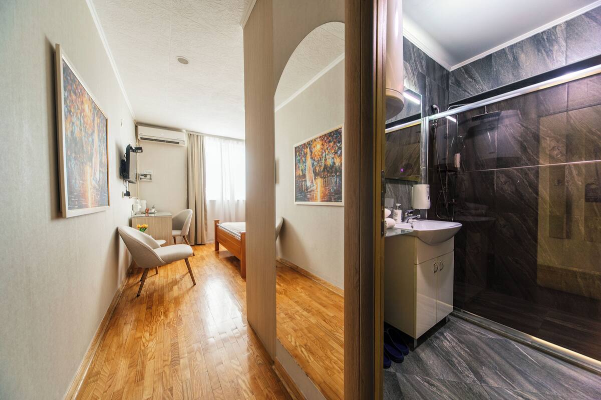 Quarto de hotel com piso de madeira, uma cama, uma escrivaninha com cadeira e banheiro com box de vidro, além de obras de arte nas paredes.