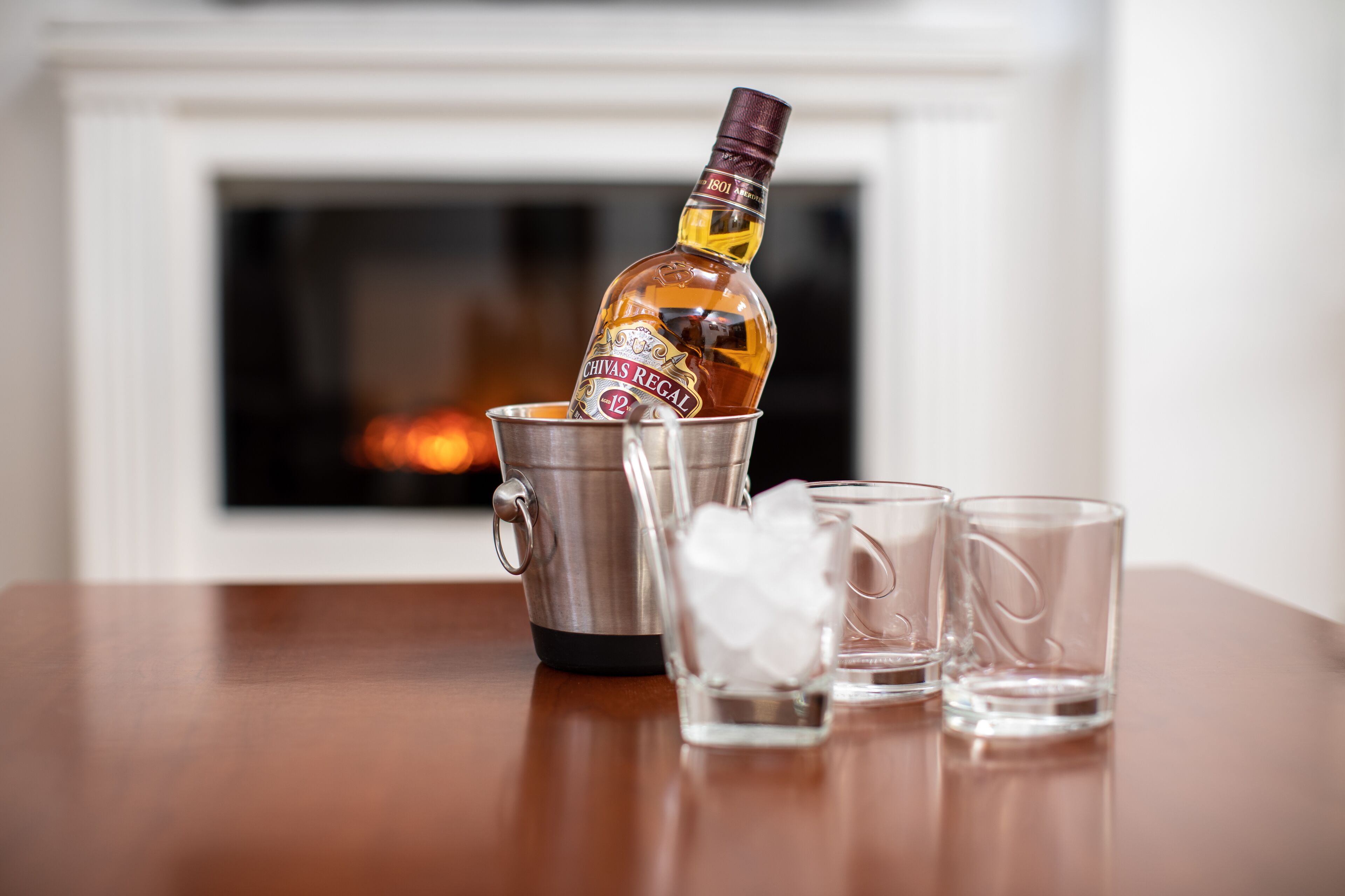A bottle of Chivas Regal whisky, a metal cup, and two shot glasses on a wooden table with a fireplace in the background.