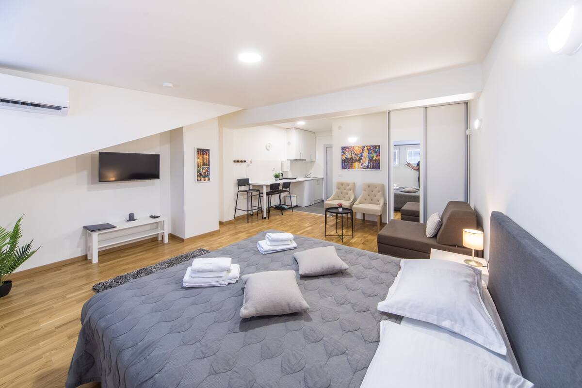 Quarto de hotel moderno com uma cama grande, uma escrivaninha, uma cadeira, um sofá e uma televisão na parede.