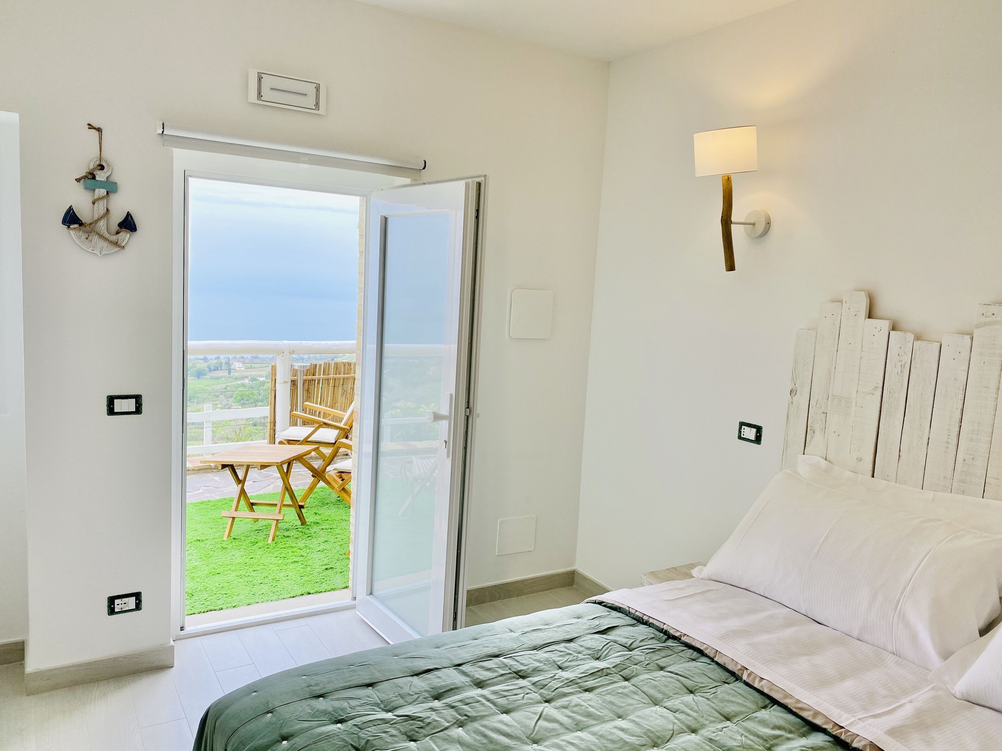 A bedroom with a bed, a nightstand, a wall-mounted lamp, and a sliding glass door leading to a balcony with a view of the sea.