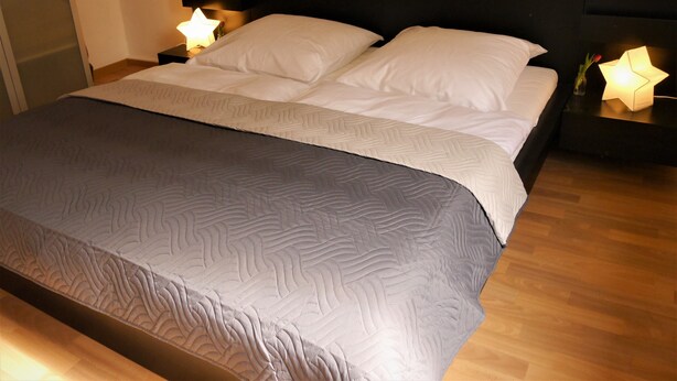 A neatly made bed with a grey patterned bedspread and white pillows, flanked by nightstands with lamps. - Eine schöne Wohnung in Kupferdreh (Essen)