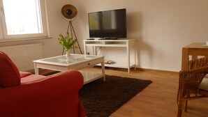 A living room with a red sofa, a glass coffee table, a TV on a white stand, a wicker chair, and a brown rug. - Eine schöne Wohnung in Kupferdreh (Essen)