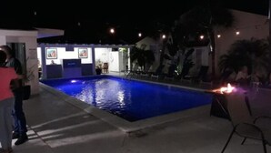 Ein Poolbereich bei Nacht mit einem blau beleuchteten Pool, Liegestühlen und einem kleinen Gebäude mit Kunstwerken.