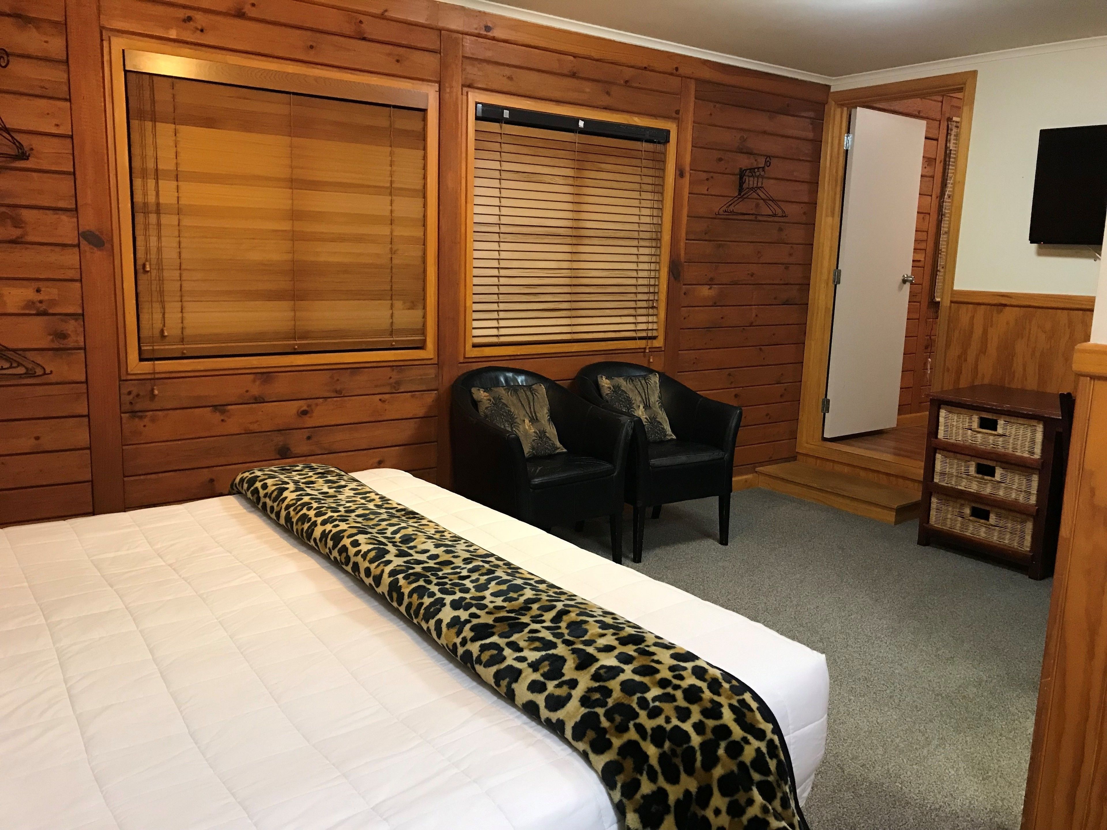 A room with wooden walls, a bed with a leopard print bedspread, two black chairs, a wooden cabinet, and a television on the wall.