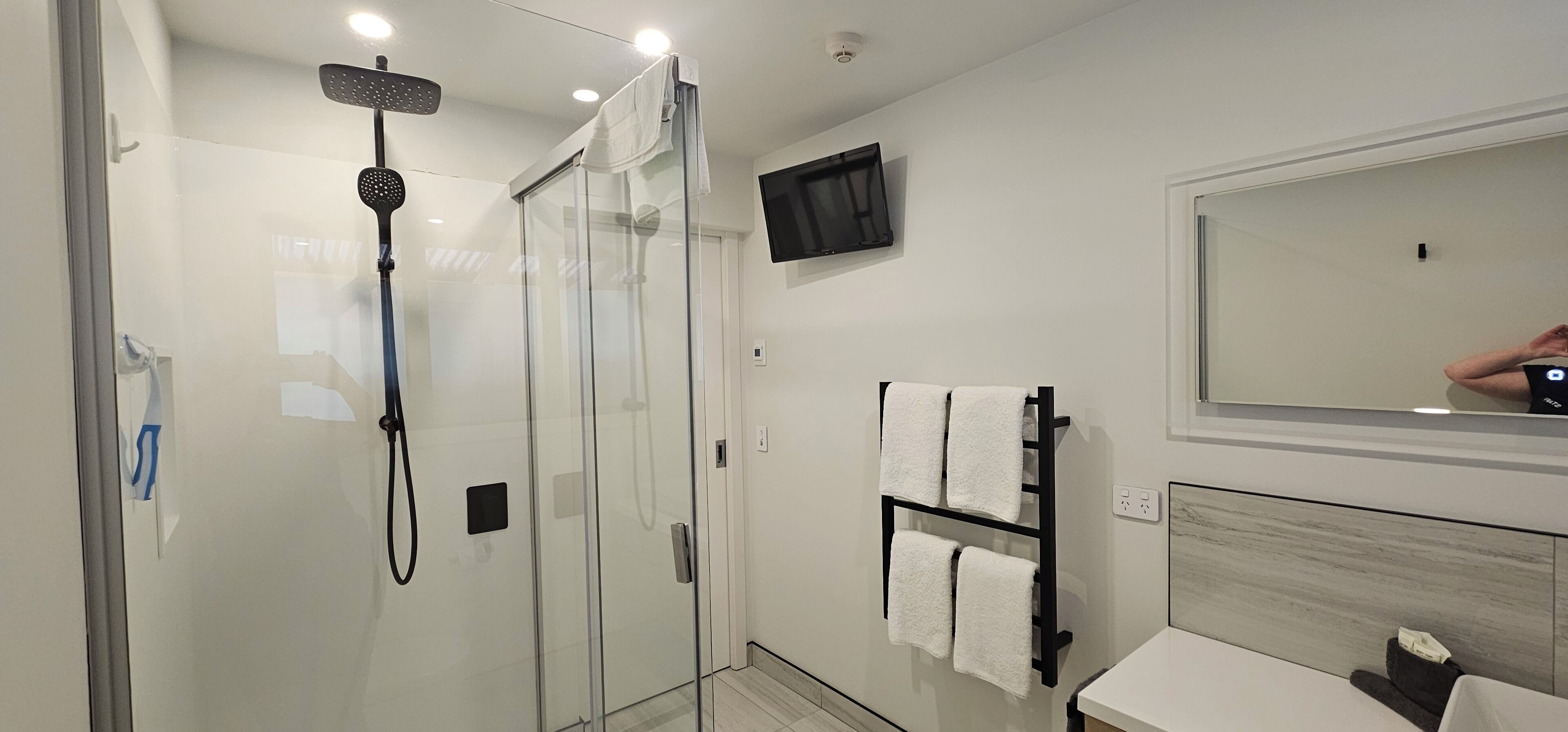 Une salle de bain moderne dotée d’une douche à parois de verre, d’un téléviseur fixé au mur et d’un porte-serviettes avec plusieurs serviettes.