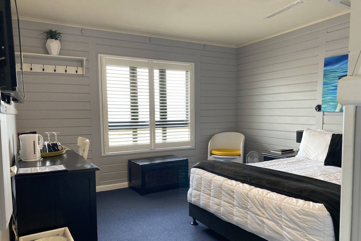 Une chambre d’hôtel comprenant un lit, un bureau avec une bouilloire, une chaise, une petite table avec un plateau, et une fenêtre avec des stores.