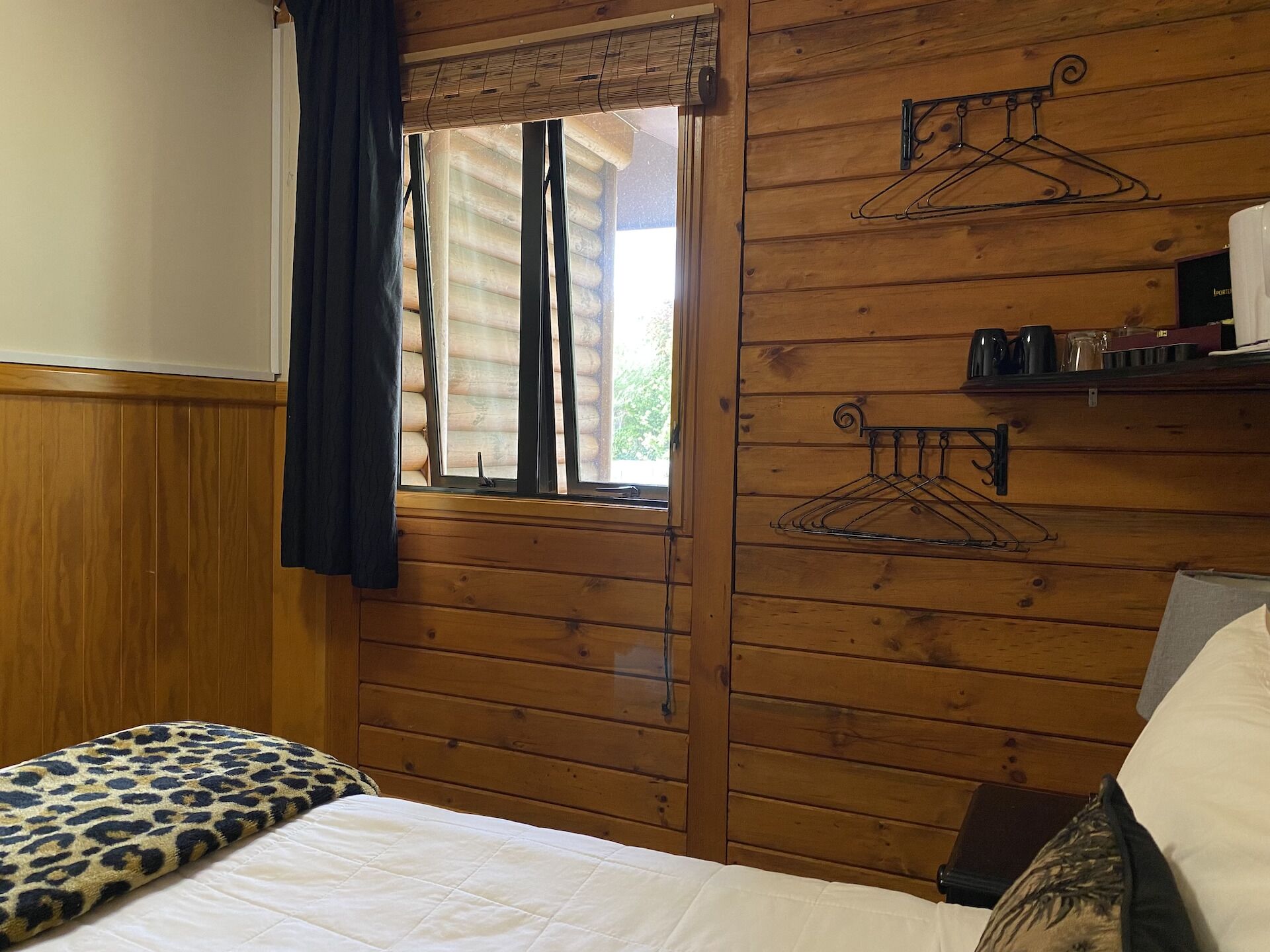 Quarto estilo cabana de madeira, com uma cama, janela com persianas e dois porta-casacos fixados na parede.