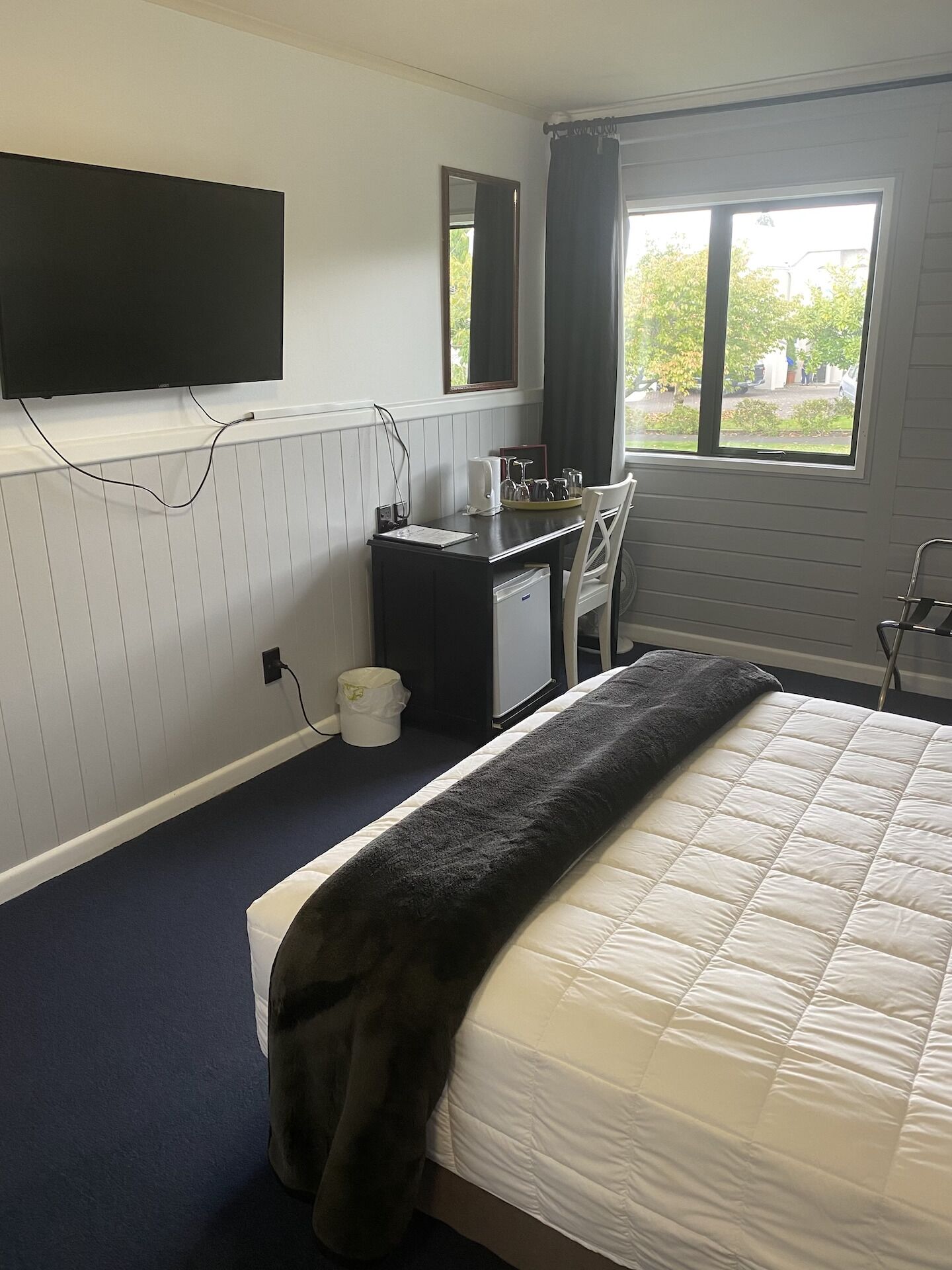 Une chambre d’hôtel comprenant un lit, un bureau avec une chaise, un téléviseur à écran plat fixé au mur, une fenêtre avec des rideaux et une vue sur l’extérieur.