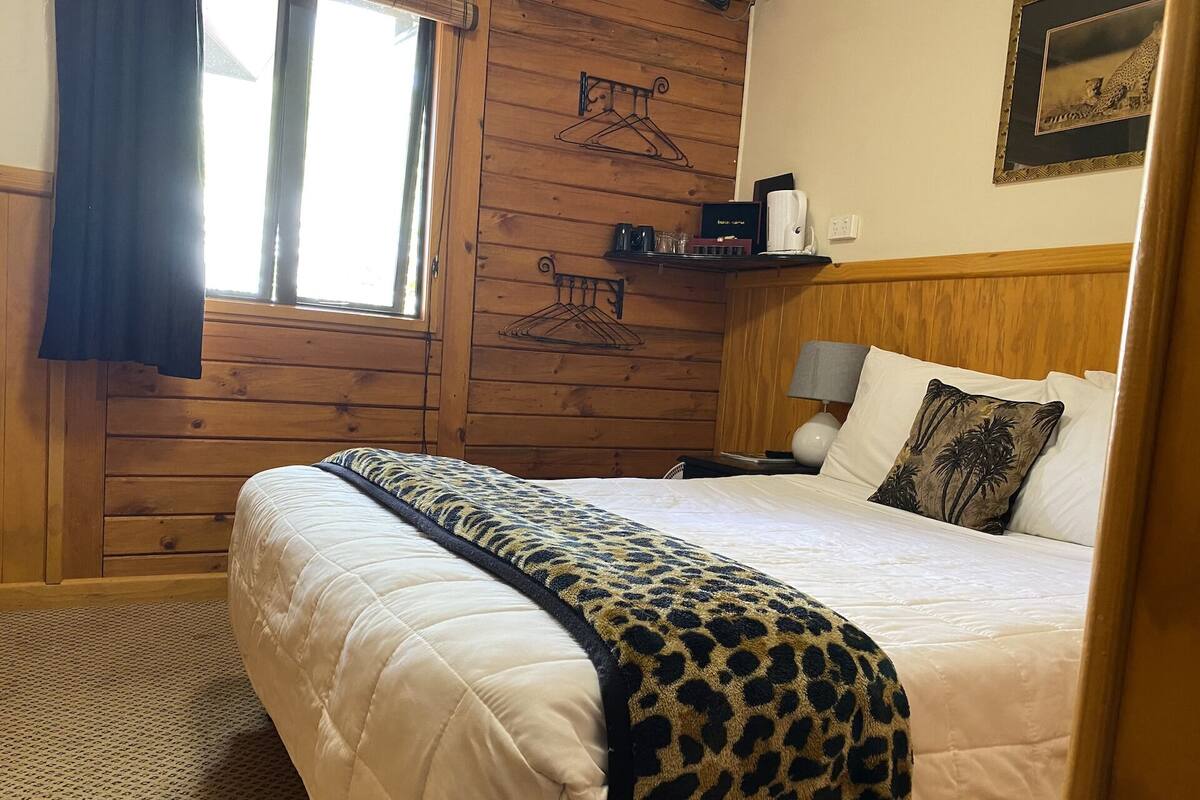 Une pièce aux murs lambrés, comprenant un lit, une table de chevet, une lampe, une fenêtre avec des rideaux et une étagère fixée au mur, sur laquelle sont posés divers objets.