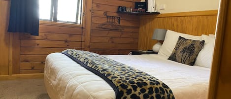 A wooden-paneled room with a bed, a nightstand, a lamp, a window with curtains, and a wall-mounted shelf with items.