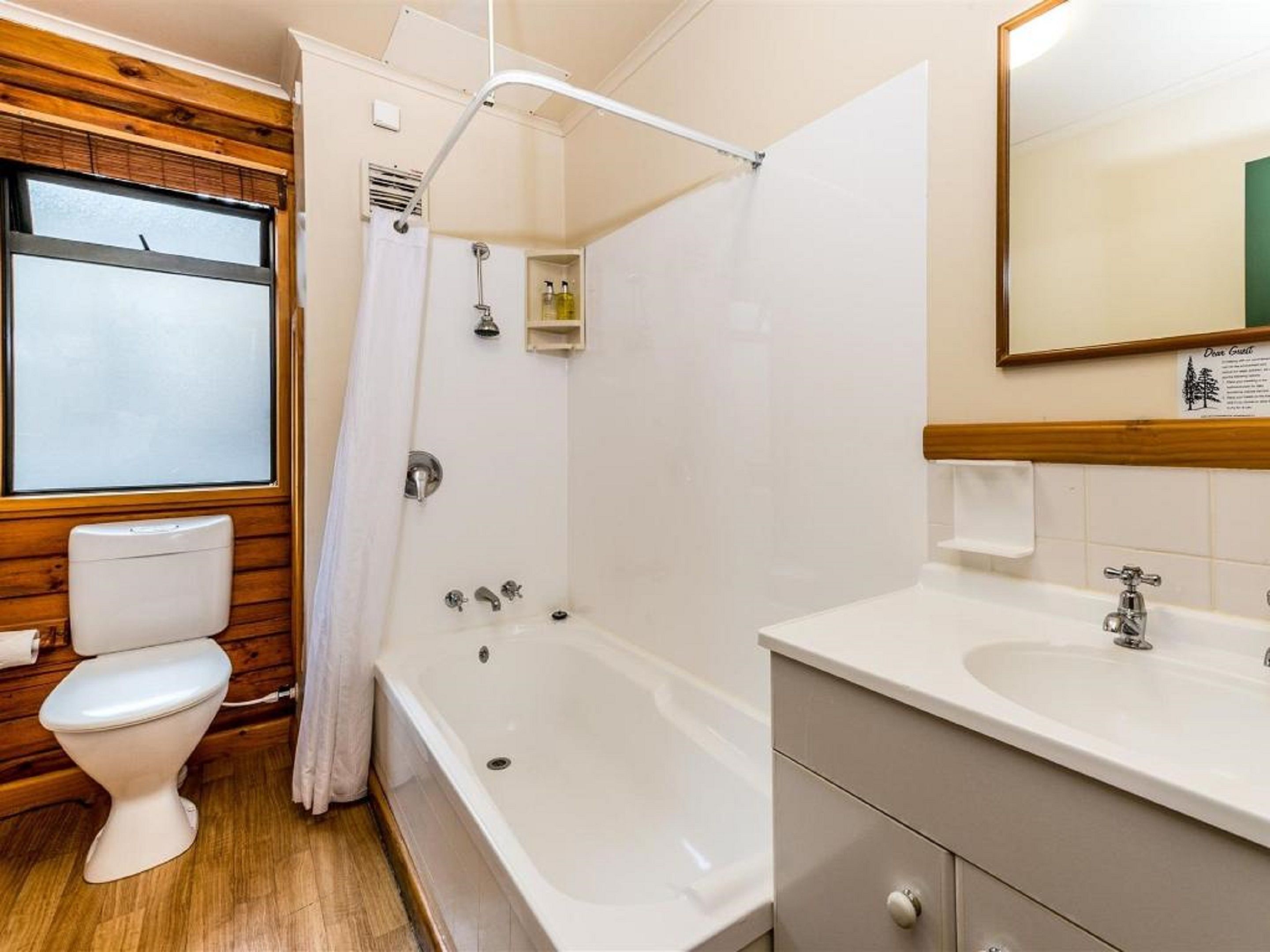 Une salle de bain avec un plancher en bois, des toilettes blanches, une baignoire et un lavabo surmonté d’un miroir.