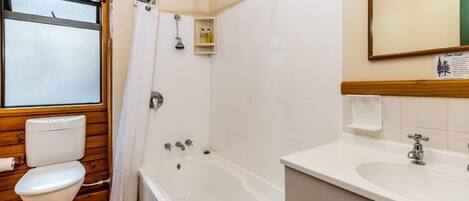 Une salle de bain avec un plancher en bois, des toilettes blanches, une baignoire et un lavabo surmonté d’un miroir.