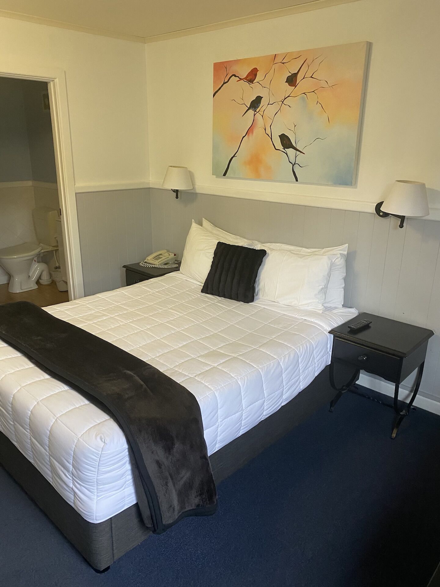 A neatly made bed with a white duvet and a dark throw blanket, a bedside table with a lamp, and a painting of birds on the wall.