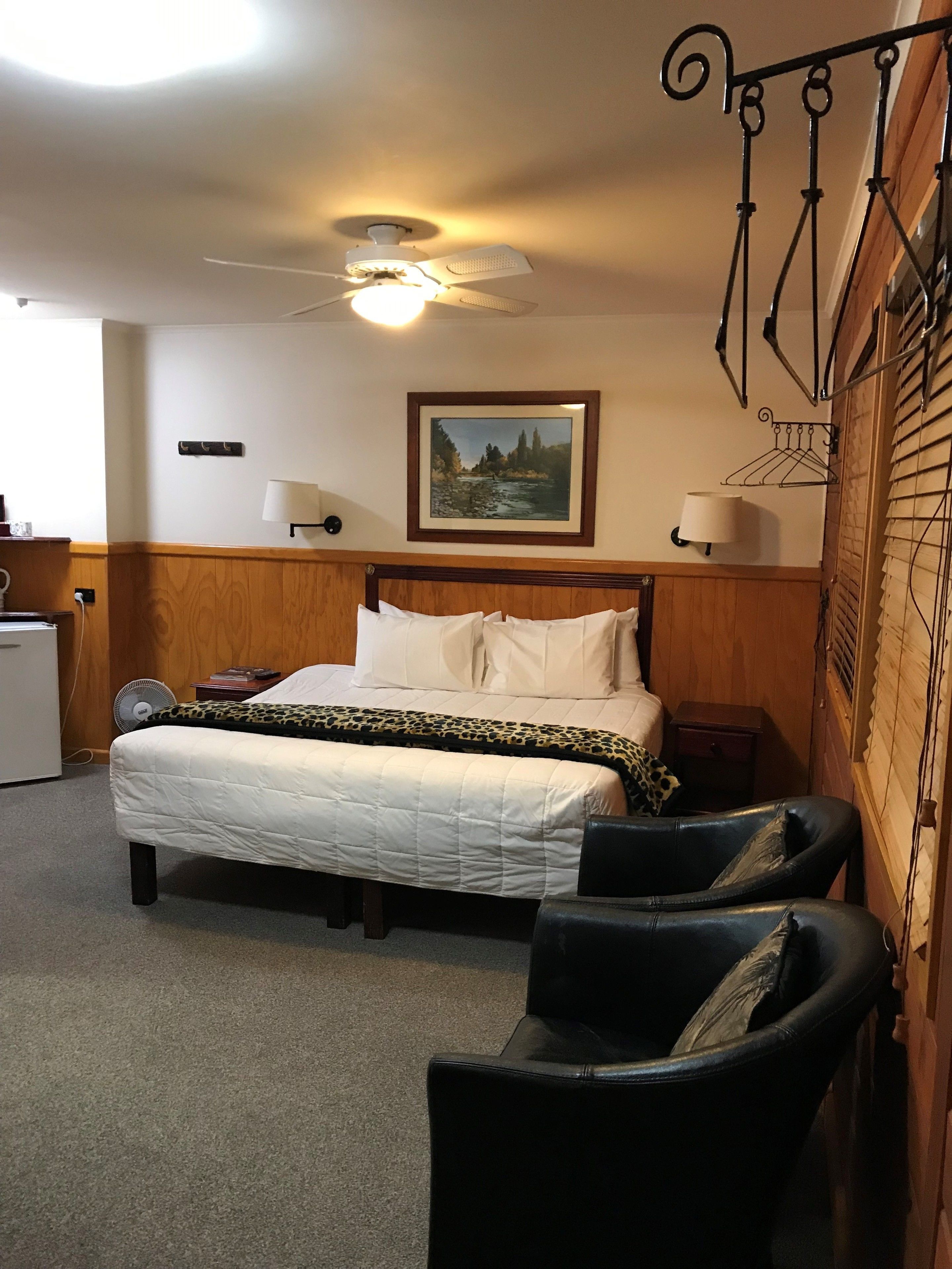 A hotel room with a bed, two chairs, a ceiling fan, a painting, and a small table.