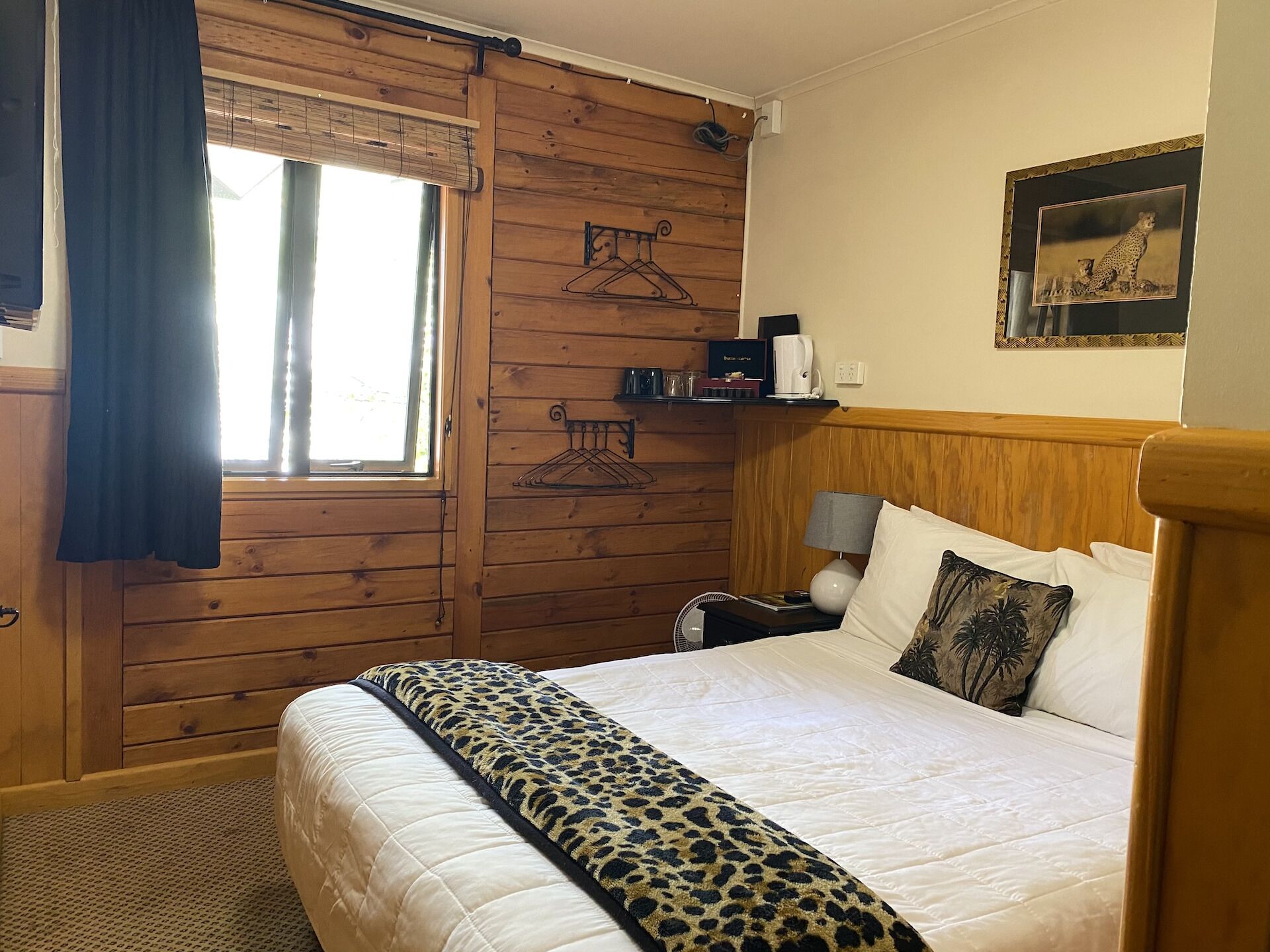 A wooden-paneled room with a bed, a window with curtains, a coat rack, a lamp, and a framed picture on the wall.