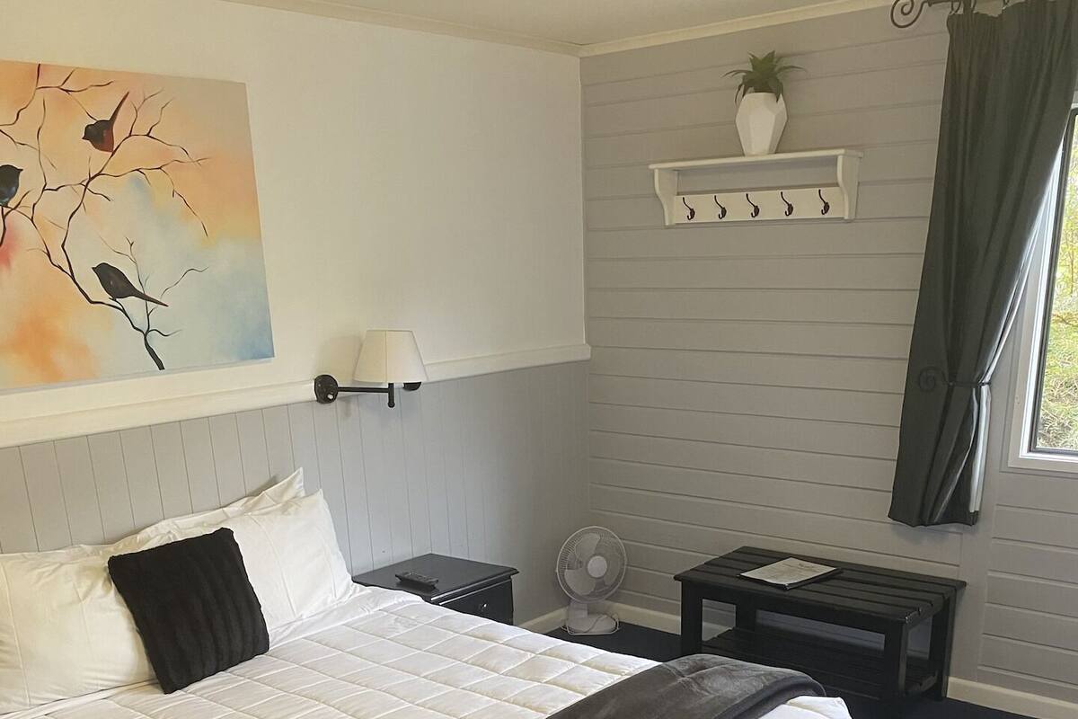 Une chambre à coucher avec un grand lit, une table de chevet avec une lampe, une petite table avec un ventilateur, une étagère avec des objets décoratifs et une fenêtre avec des rideaux.