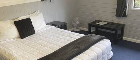 Un dormitorio con una cama grande, una mesita de noche con lámpara, una pequeña mesa con ventilador, un estante con objetos decorativos y una ventana con cortinas.