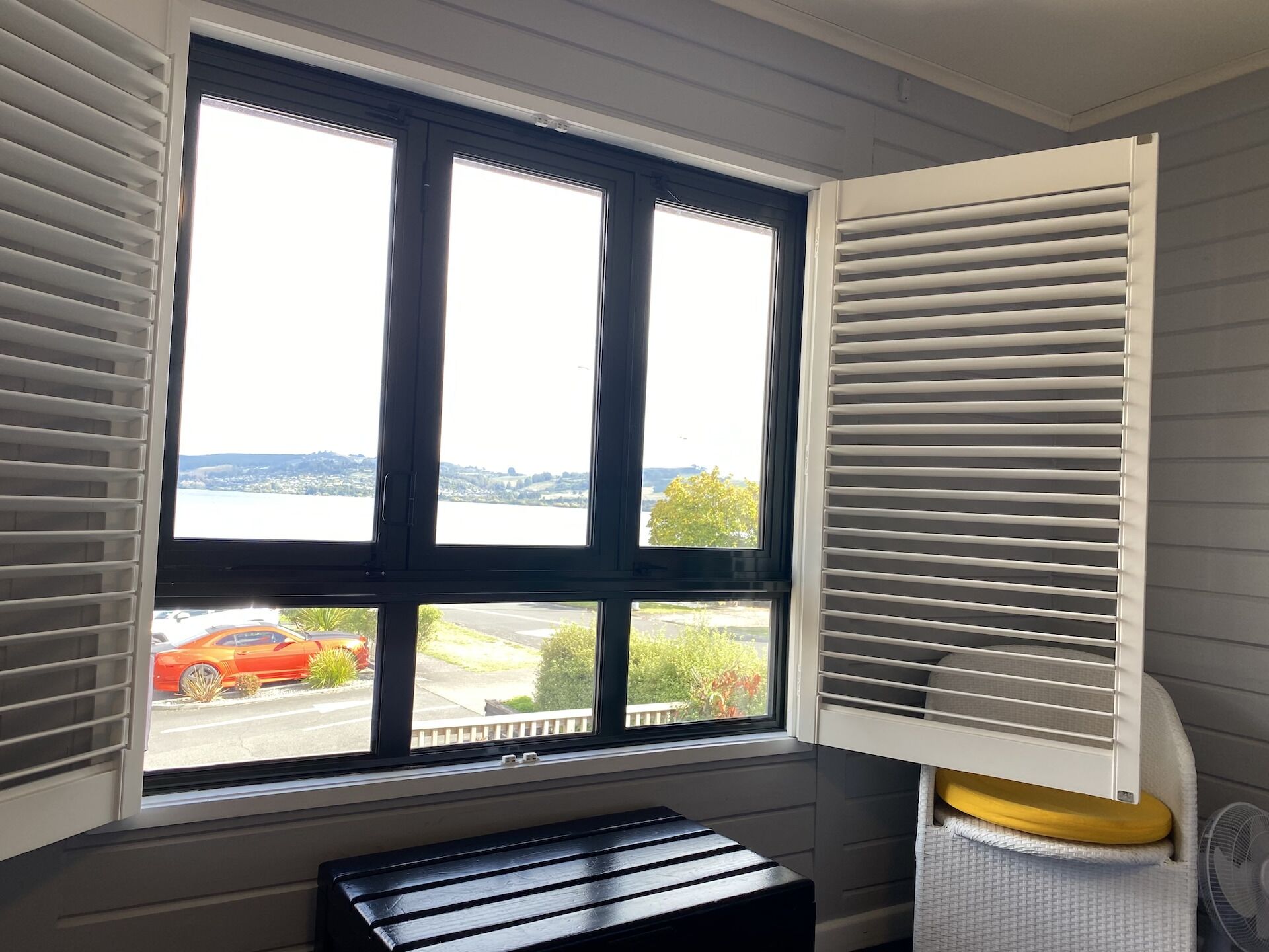 A room with a large window, white blinds, a black bench, and a white fan.