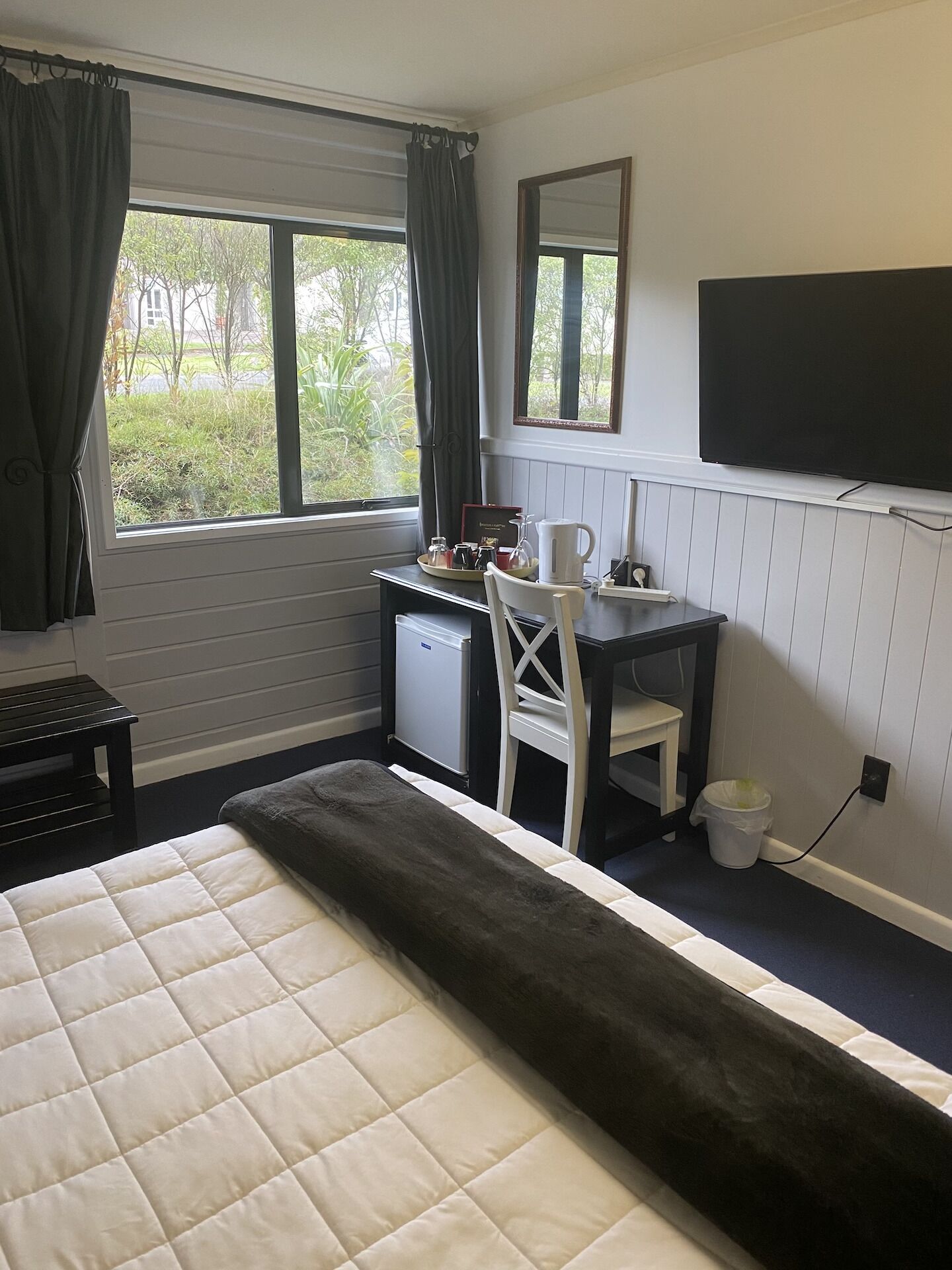 Habitación de hotel con cama, escritorio y silla, refrigerador pequeño, televisor y ventana con cortinas.