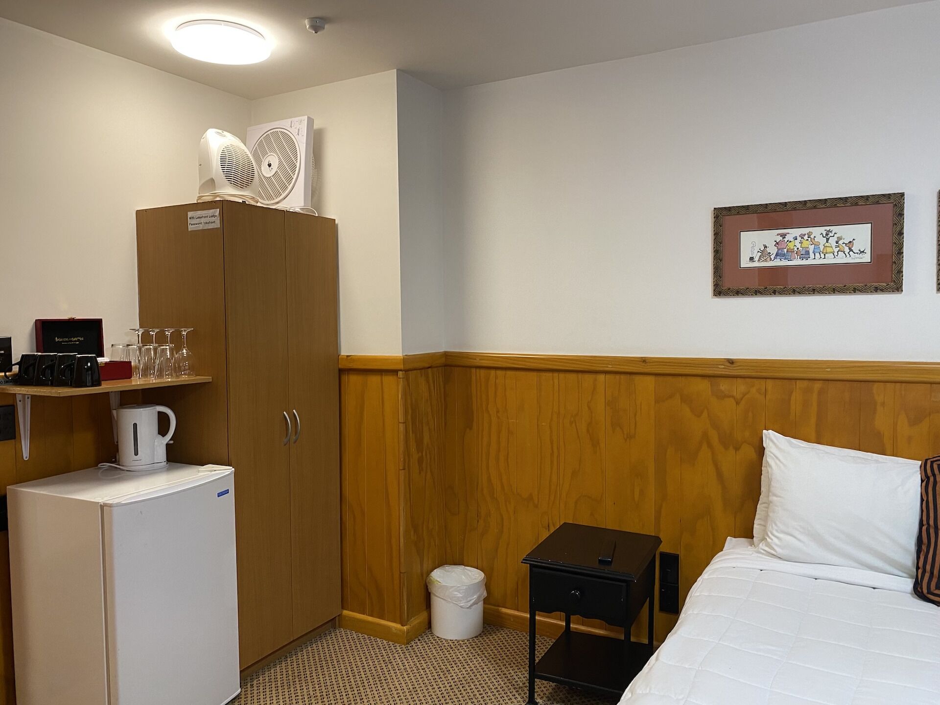Ein Hotelzimmer mit Bett, einem kleinen Nachttisch, einem Schrank, einem Wasserkocher und einem Regal mit Gläsern und einer Kaffeemaschine.