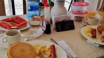 A breakfast table with plates of pancakes, scrambled eggs, and a hot dog, along with a bottle of water, a coffee cup, and a pitcher of orange juice.