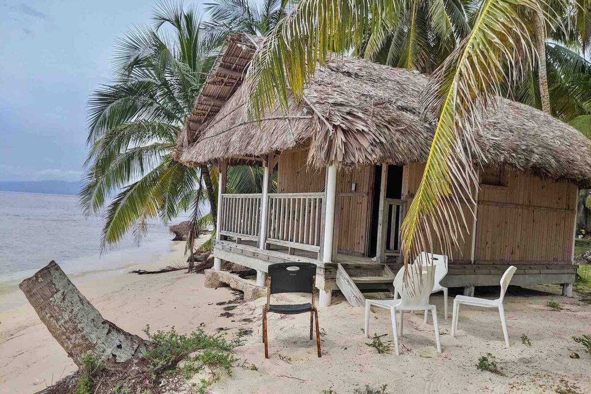 A beach hut with a thatched roof, white railings, and a small porch area. There are two white plastic chairs and a black chair outside.