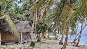 A beach scene with palm trees, a thatched-roof hut, and a hammock. -  San Blas Paradise Private Cabins on Shipwreck Island - meals included (Ailigandi)